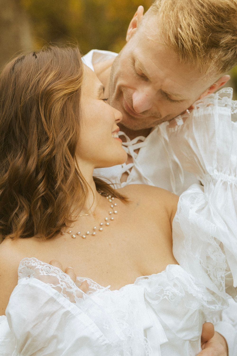 Close-up of bride in white lace gown and pearl necklace sharing a kiss with groom outdoors.