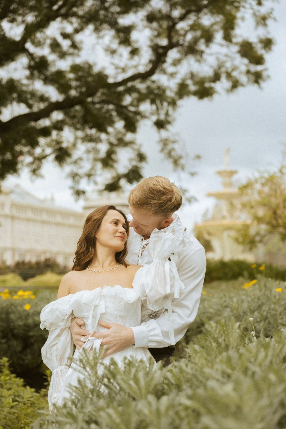 A romantic couple in white period attire embrace in a lush garden with a fountain and grand building backdrop.
