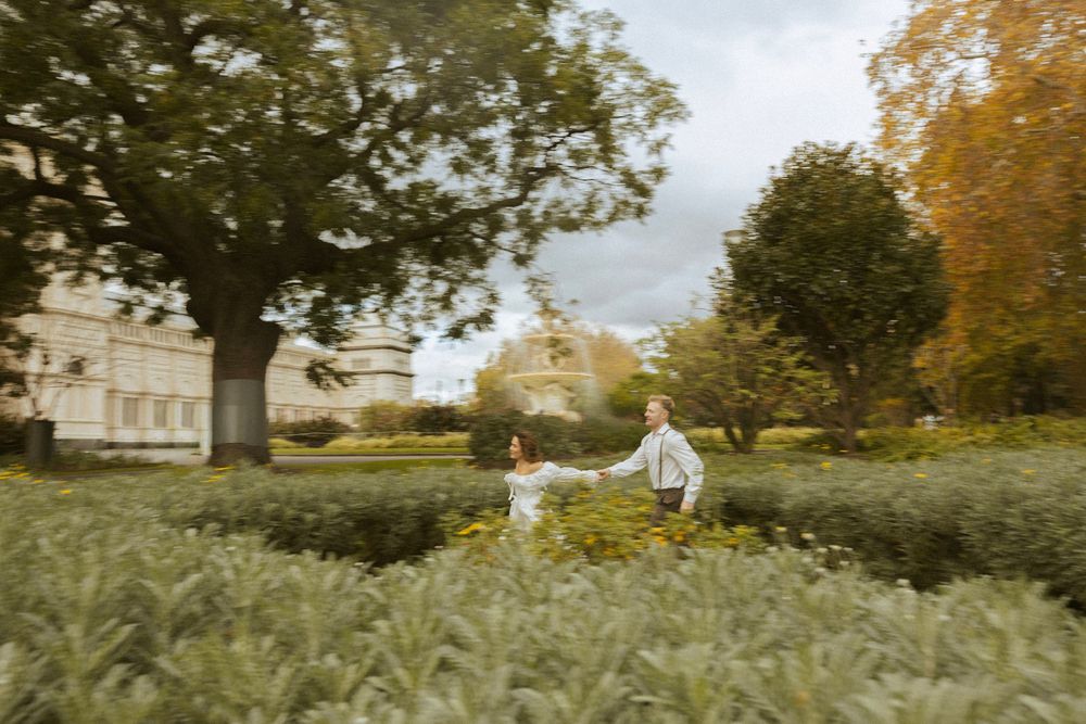 Bride and groom holding hands running through green garden path with historic building behind.