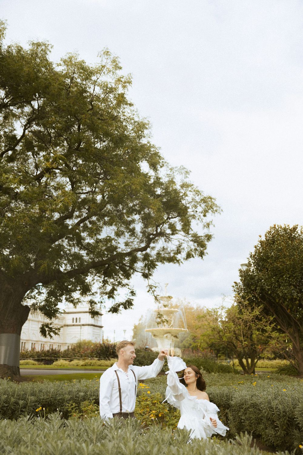 Bride and groom dancing under large tree near fountain in lush garden.