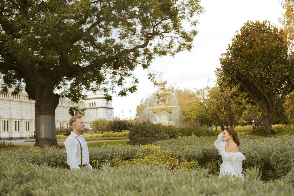 Bride and groom standing apart in a sunlit garden with a grand estate building in background.