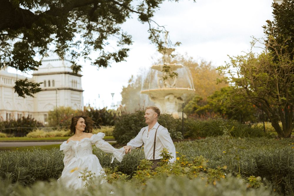 Couple holding hands in wildflower garden with ornate fountain and building behind.