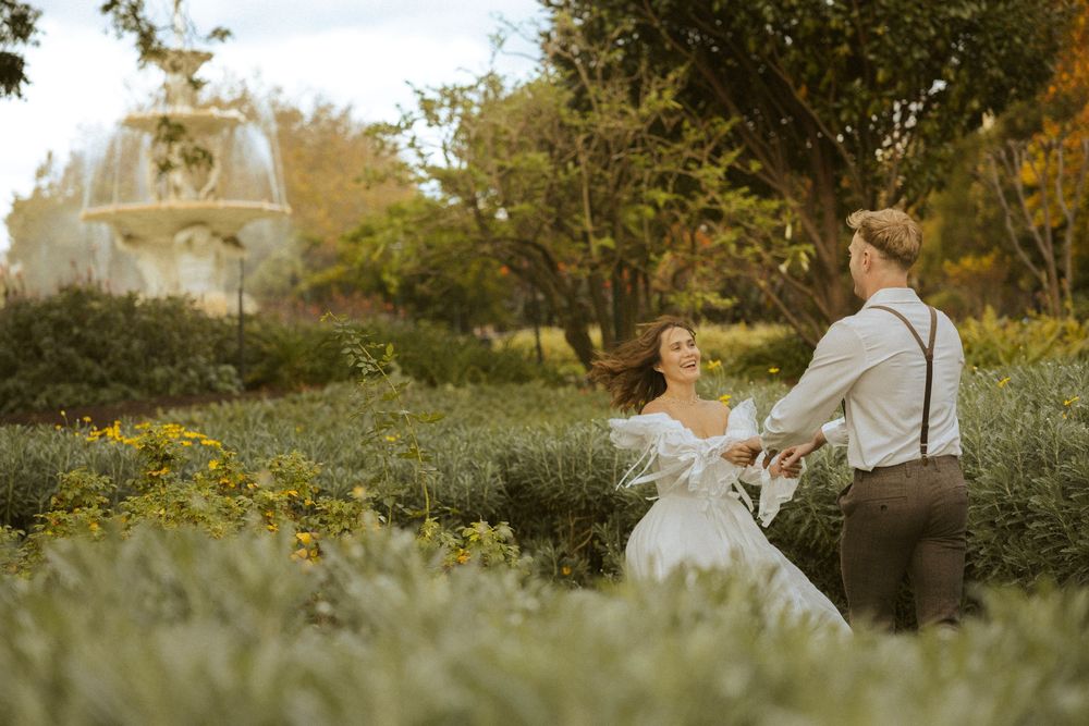 Couple dancing joyfully in blooming garden with stone fountain, bride in elegant white dress.