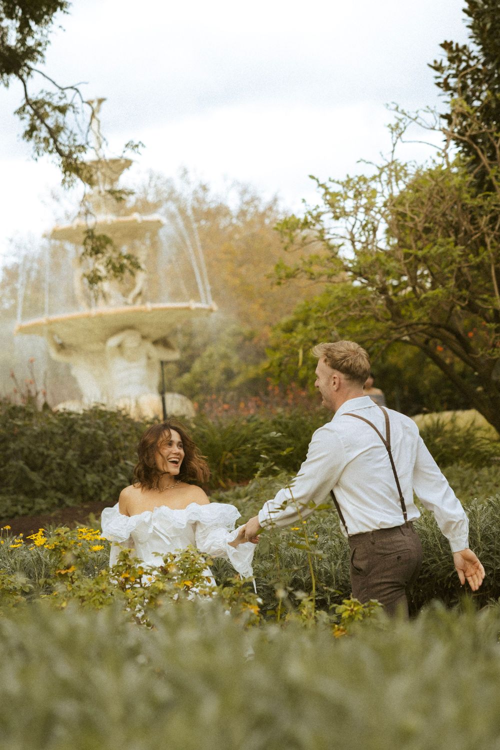 Joyful couple holding hands and laughing near a stone fountain surrounded by lush greenery.