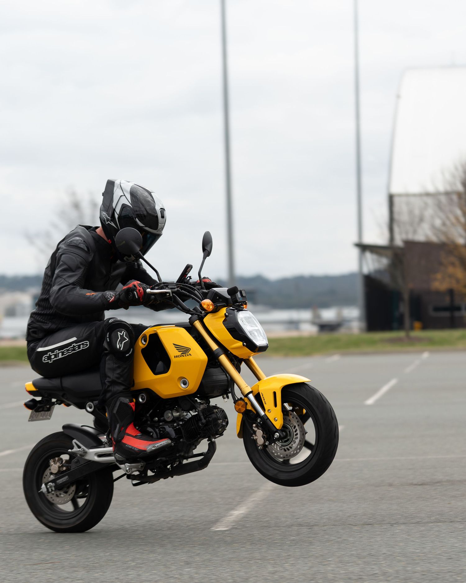 Rider performing a wheelie on a yellow Honda Grom mini motorcycle in a parking lot.