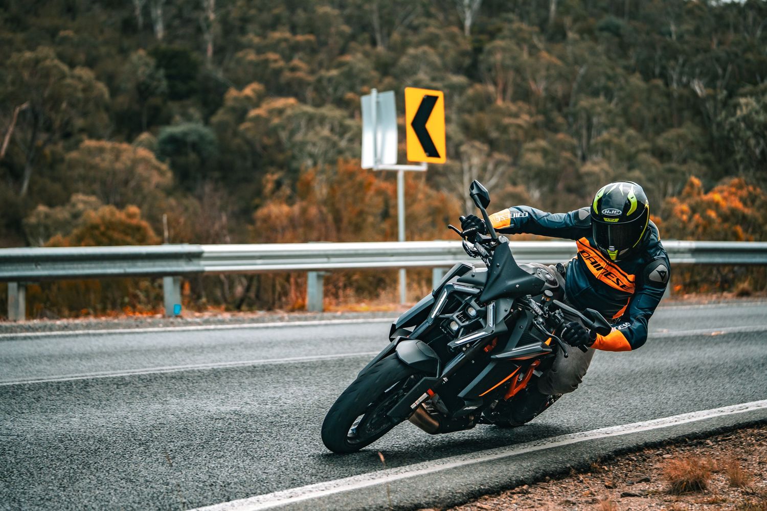 Rider leaning a black KTM Duke motorcycle into a wet curve on an autumn road.