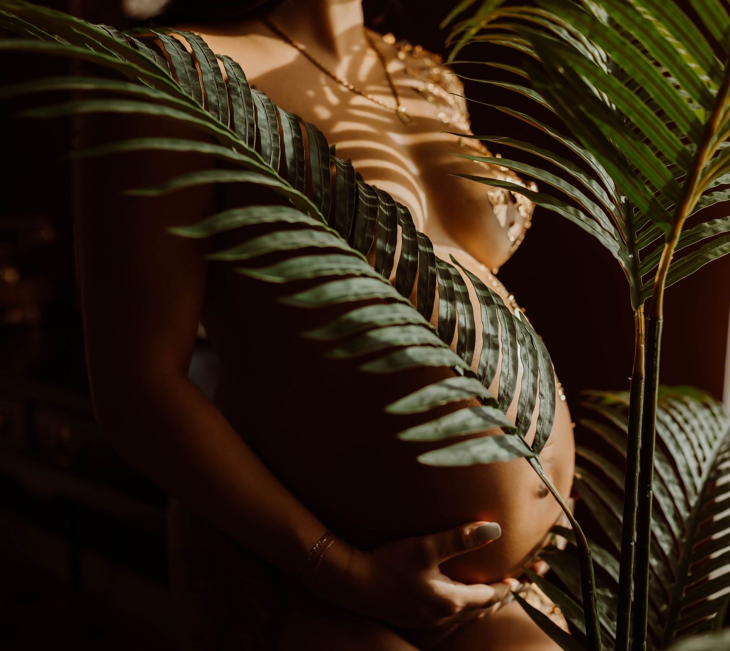 A pregnant woman cradles her baby bump surrounded by lush tropical palm leaves in warm golden light - Stillwater boudoir photographer.