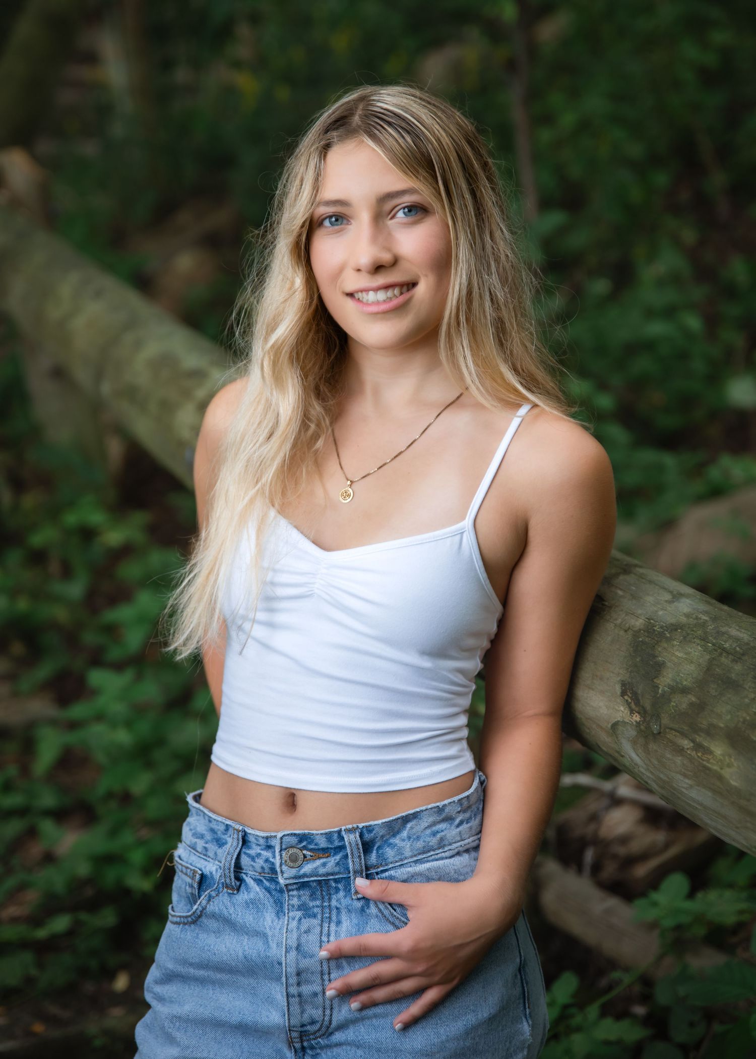 Young blonde woman in white crop top and jeans poses outdoors against a fallen log surrounded by lush greenery.
