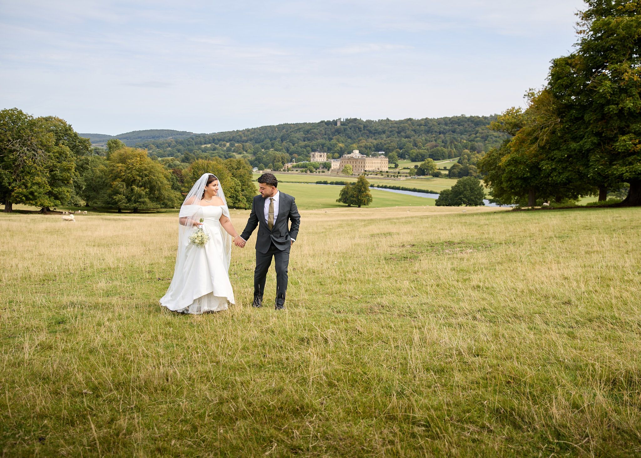 Bride and groom walking hand in hand through a green meadow with a grand estate in the background.