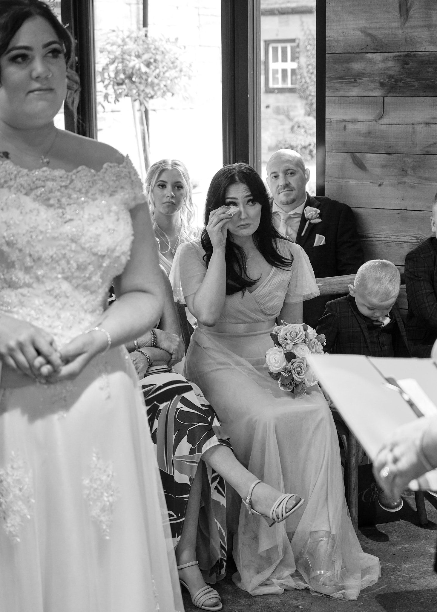 Emotional bridesmaid wiping tears during wedding ceremony in black and white candid photo.