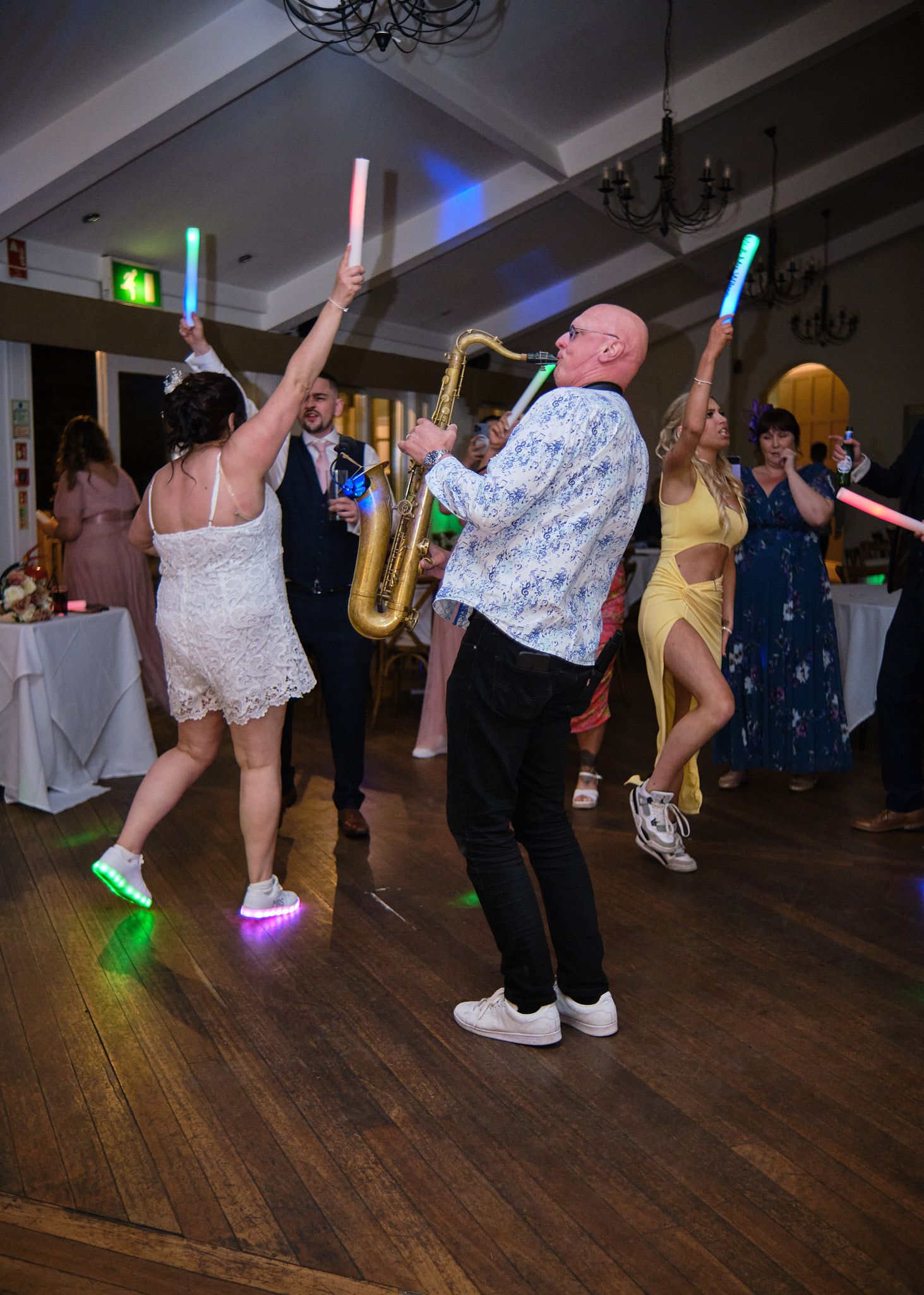 Wedding guests dancing with glowing foam sticks while saxophonist performs at lively reception.