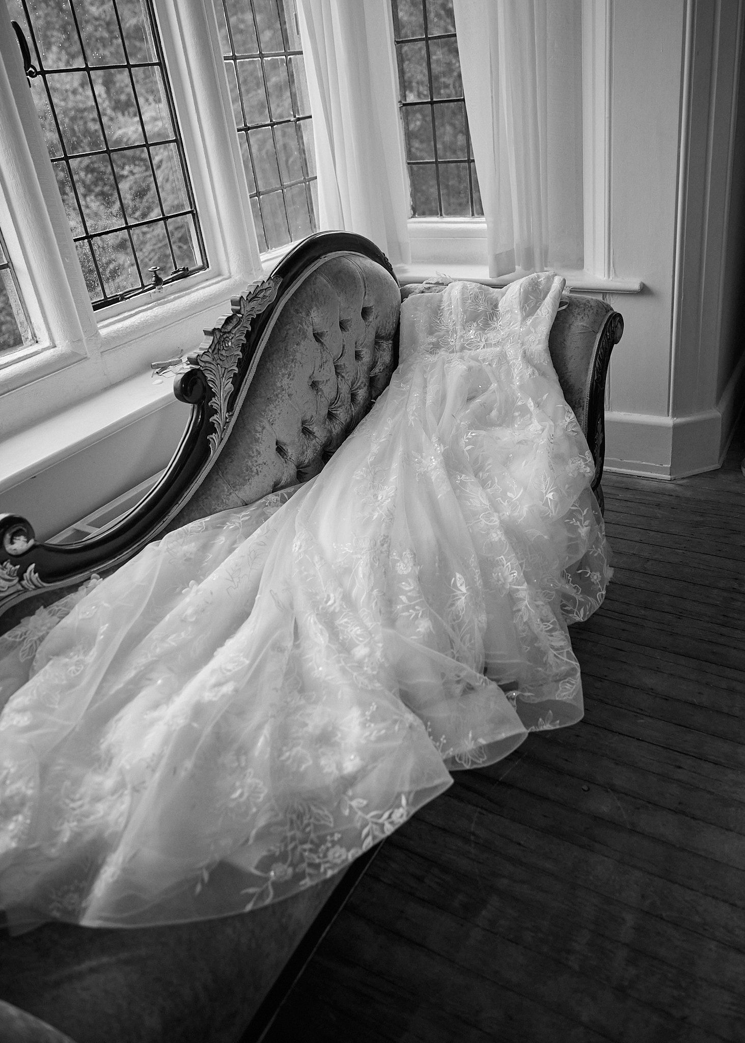 Black and white photo of an elegant lace wedding dress draped over a vintage tufted chaise lounge near sunlit windows.