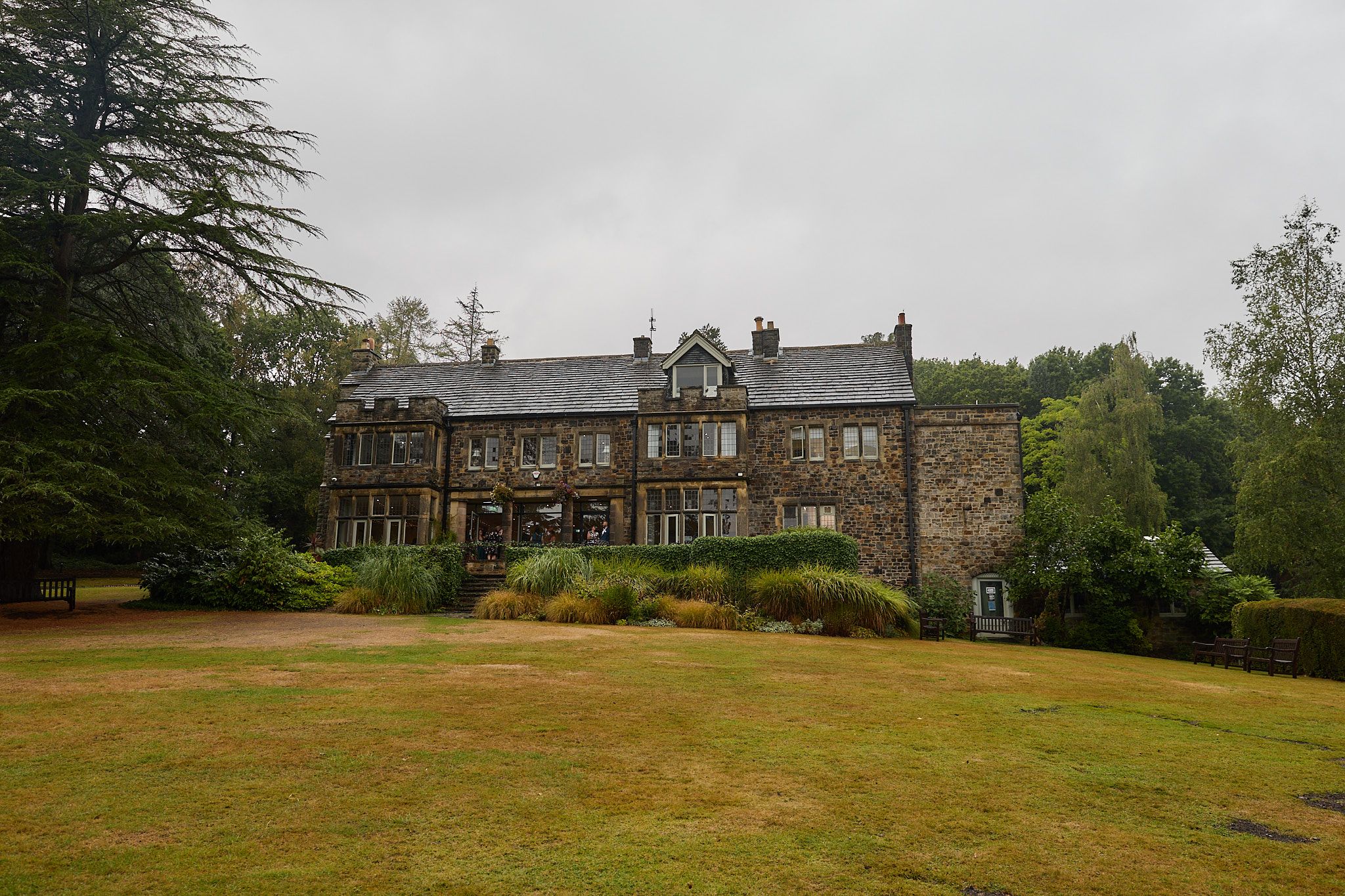 Historic stone manor house surrounded by lush green lawns and mature trees under an overcast sky.