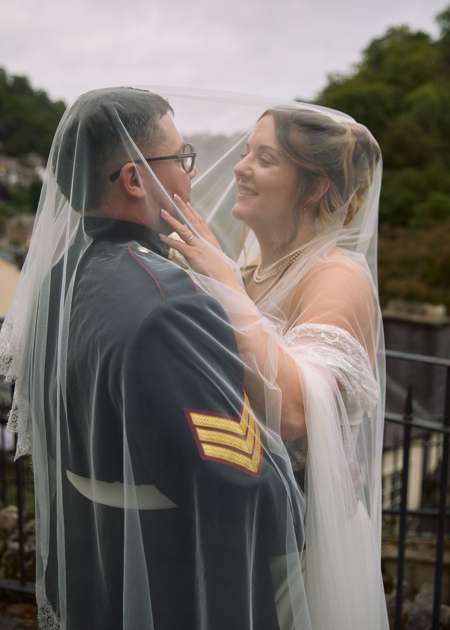 Bride and groom share romantic moment under wedding veil, groom in military dress uniform with sergeant stripes.