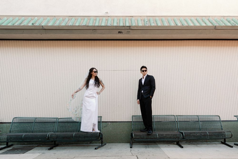 Bride in white gown and groom in black suit posing apart on benches outside a building.