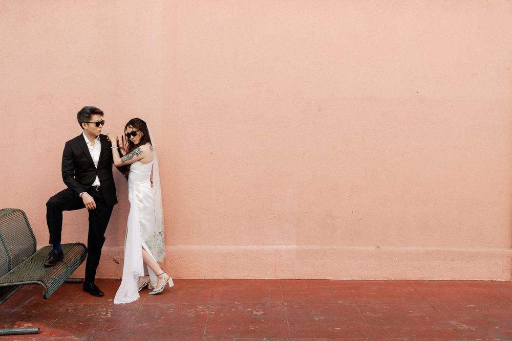 Stylish couple in sunglasses posing against a pink wall, man in black suit and woman in white dress.