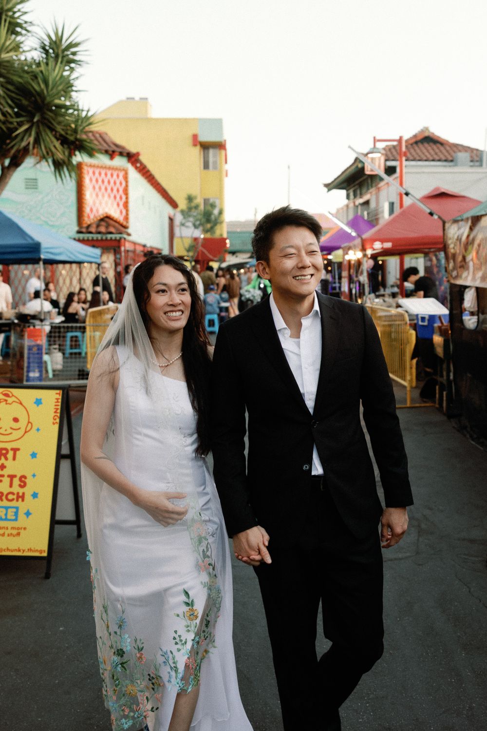 Asian couple in wedding attire walking hand-in-hand at a colorful outdoor market, bride in floral white gown with veil.