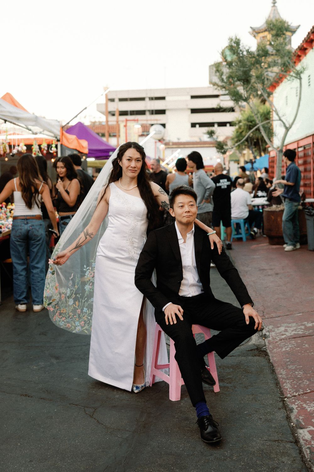 A bride in a white gown and groom in a black suit pose at an outdoor market in Chinatown.