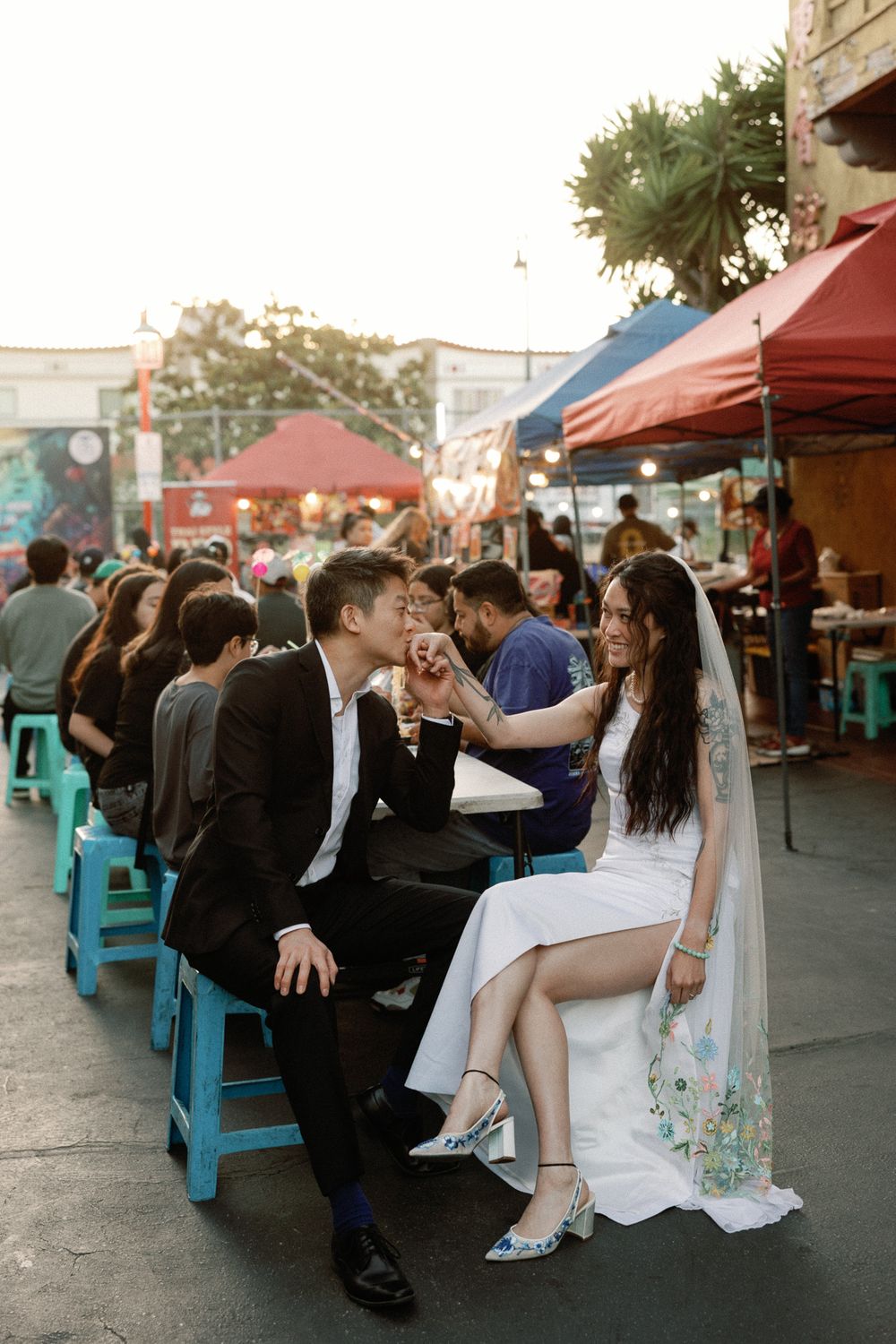Bride and groom share a romantic moment at a vibrant outdoor market, she feeds him while wearing a white gown.