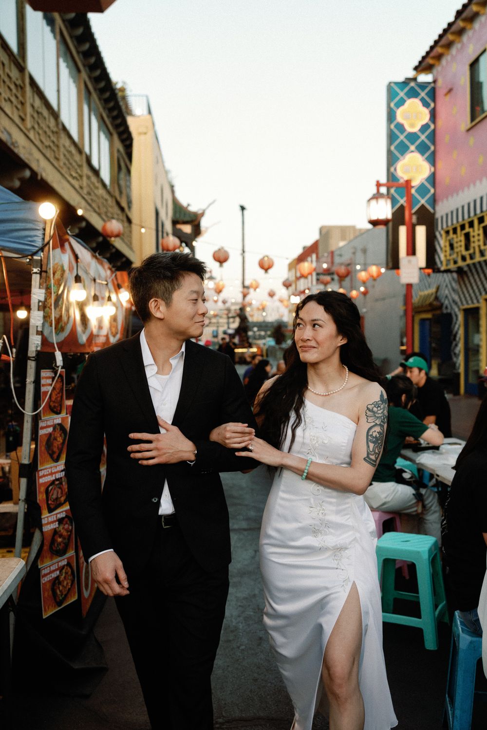 Couple in wedding attire walking through Chinatown street market with lanterns at dusk.
