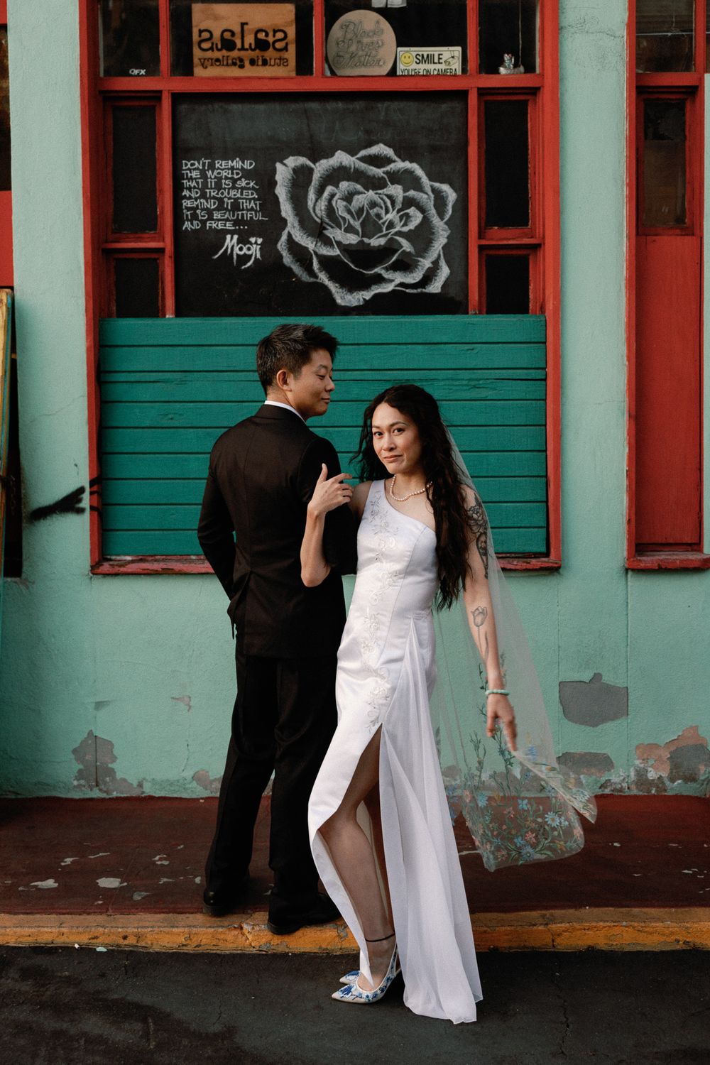 Asian wedding couple posing in front of a turquoise building with rose mural, bride in white slit gown with tattoos.