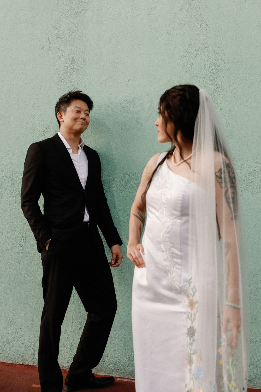 Groom in black suit smiling at bride in embroidered wedding dress with veil by mint wall.