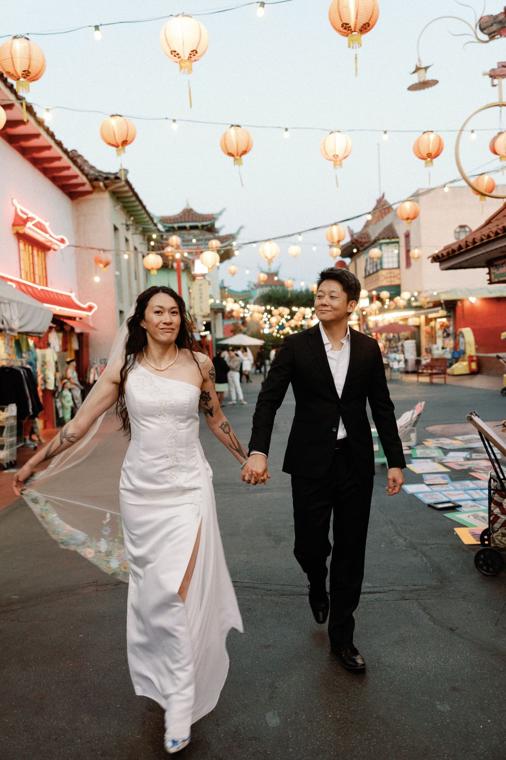 A couple in wedding attire walks hand-in-hand through a vibrant Chinatown street adorned with glowing red lanterns at dusk.