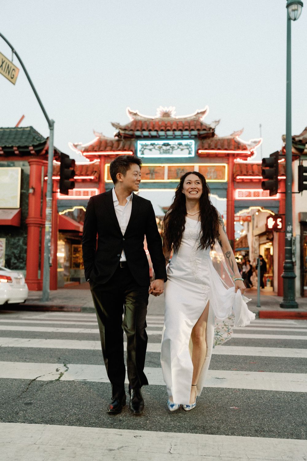 Newlywed couple holds hands crossing street in front of illuminated Chinatown gate at dusk.