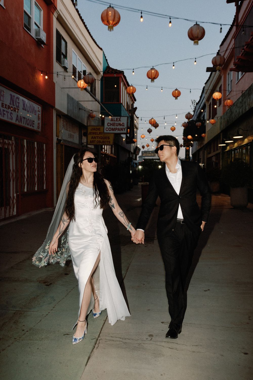 Newlyweds holding hands walking under red lanterns in Chinatown street at twilight.