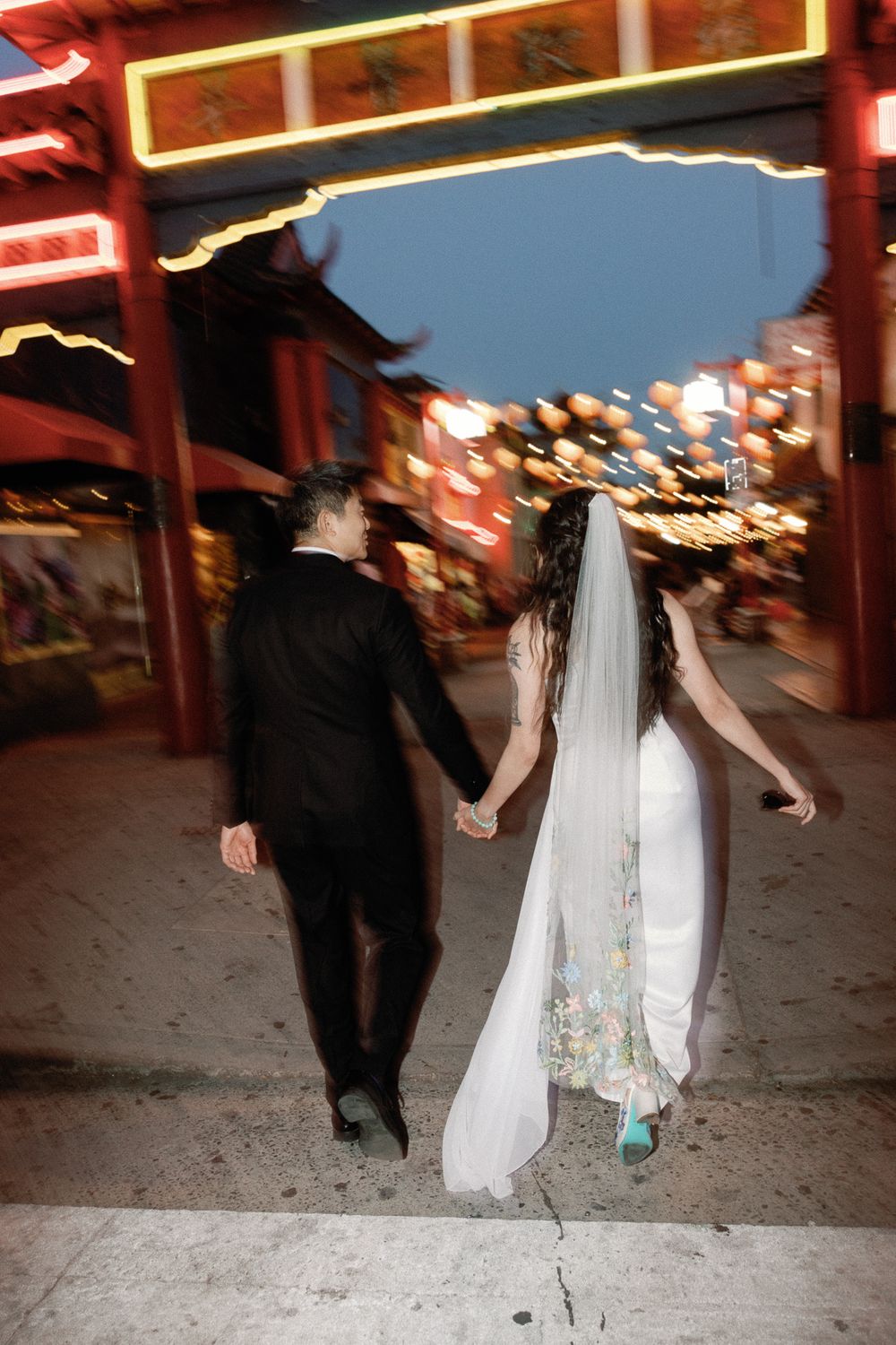 Bride and groom walk hand-in-hand through a vibrant Chinatown at night, surrounded by glowing lights and red archways.