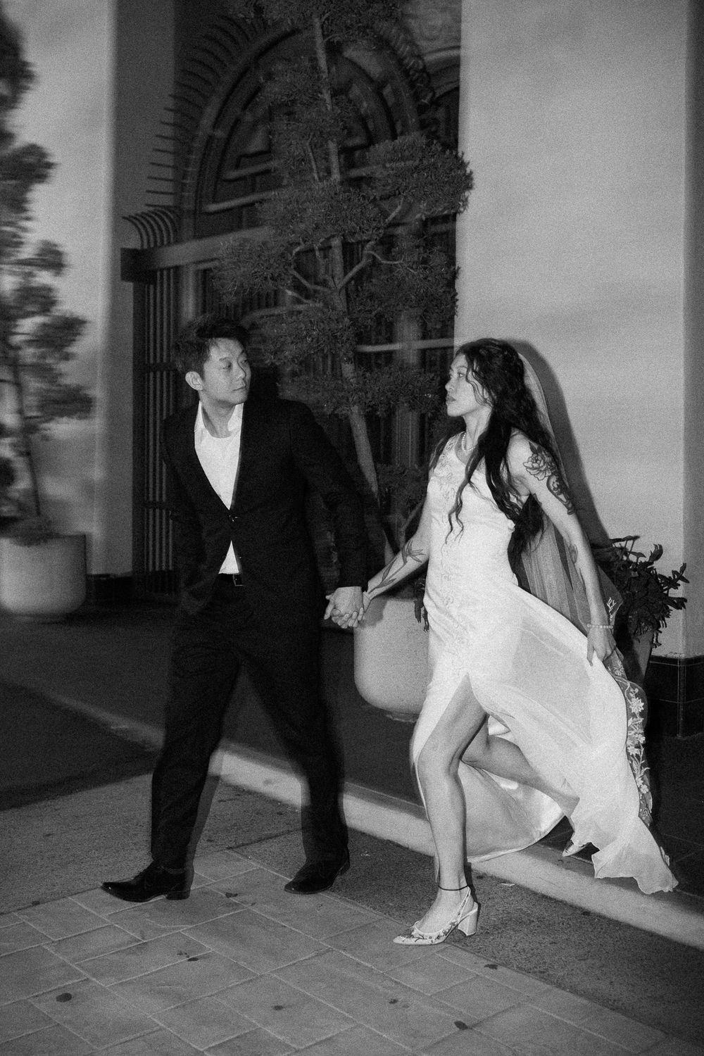 Black and white photo of a couple in wedding attire holding hands and walking outside a building.