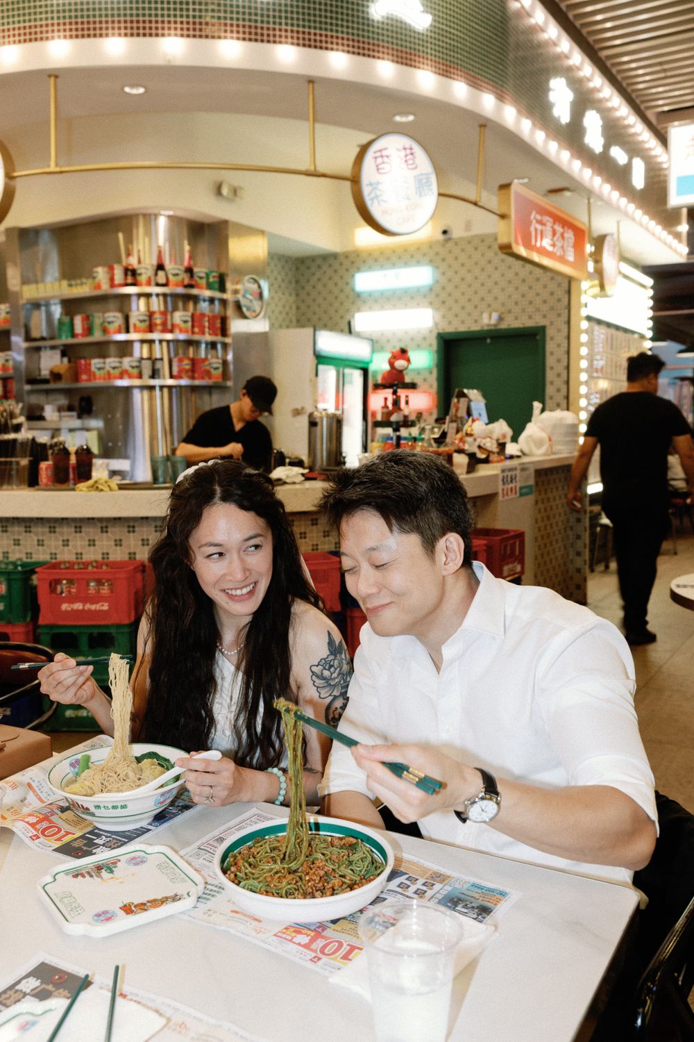 Couple enjoying noodle bowls at an Asian restaurant, smiling and using chopsticks in a vibrant food court setting.