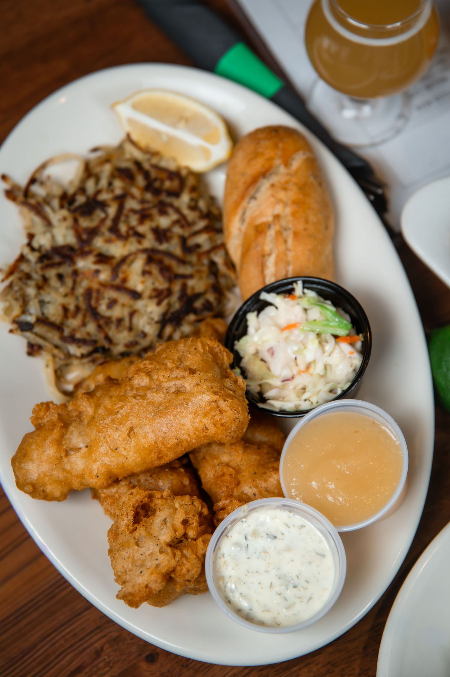 A plate of golden fried fish, hash browns, coleslaw, a bread roll, and dipping sauces with a lemon wedge.