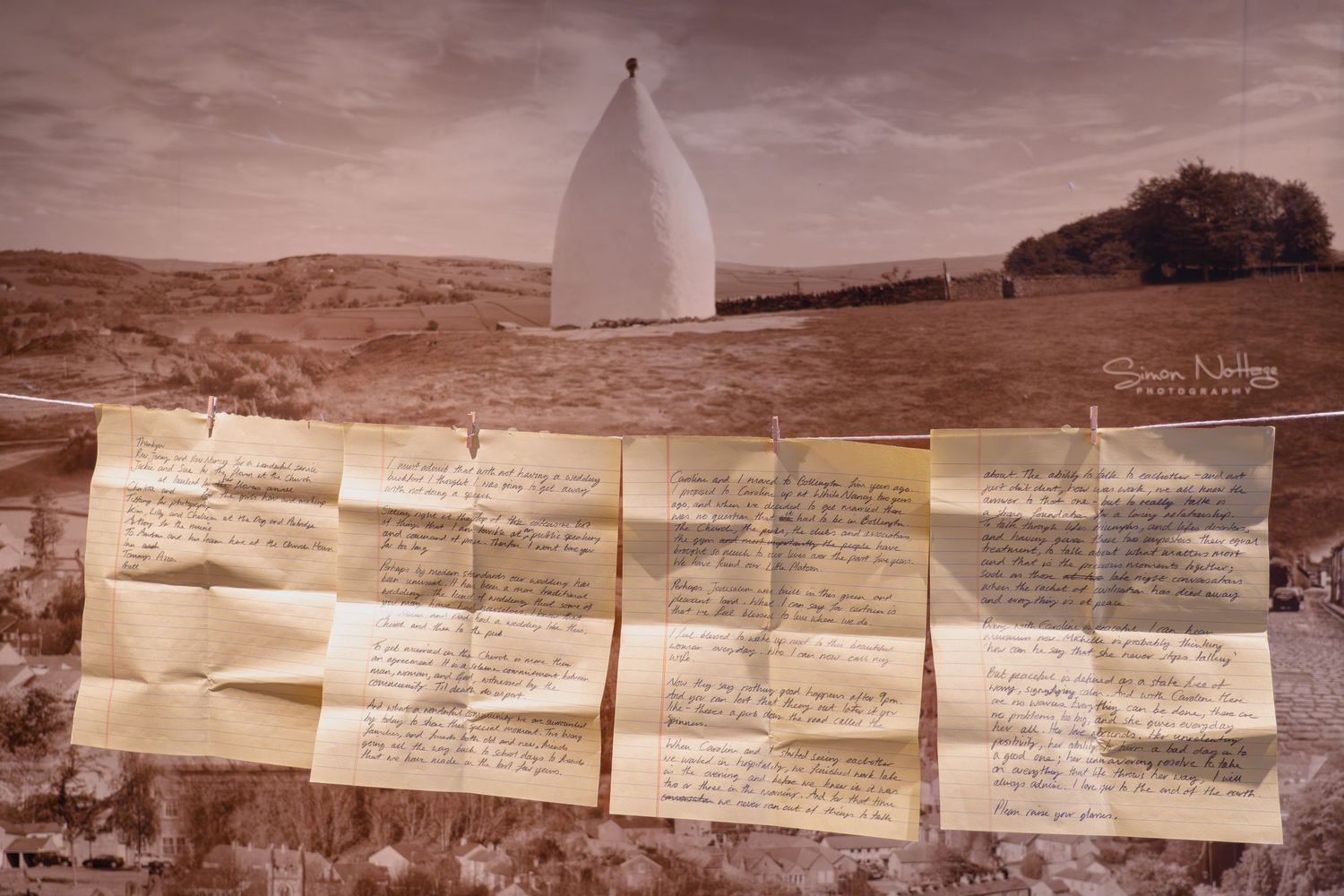 Pages from the groom's wedding speech clipped to a line against a scenic landscape photograph, creating a documentary-style wedding portrait