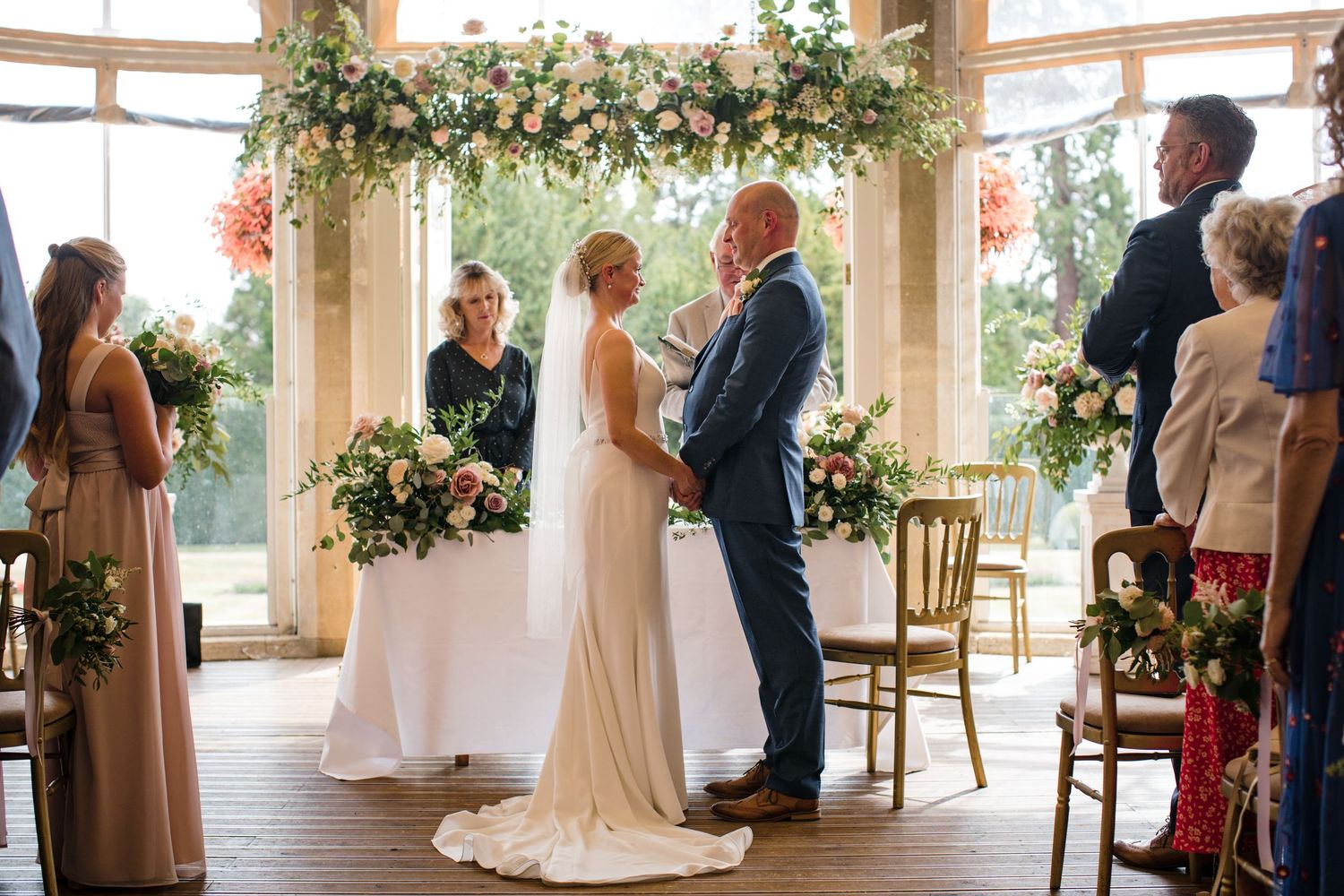 Couple exchanging vows at elegant indoor wedding ceremony with floral arch and pink flower arrangements.