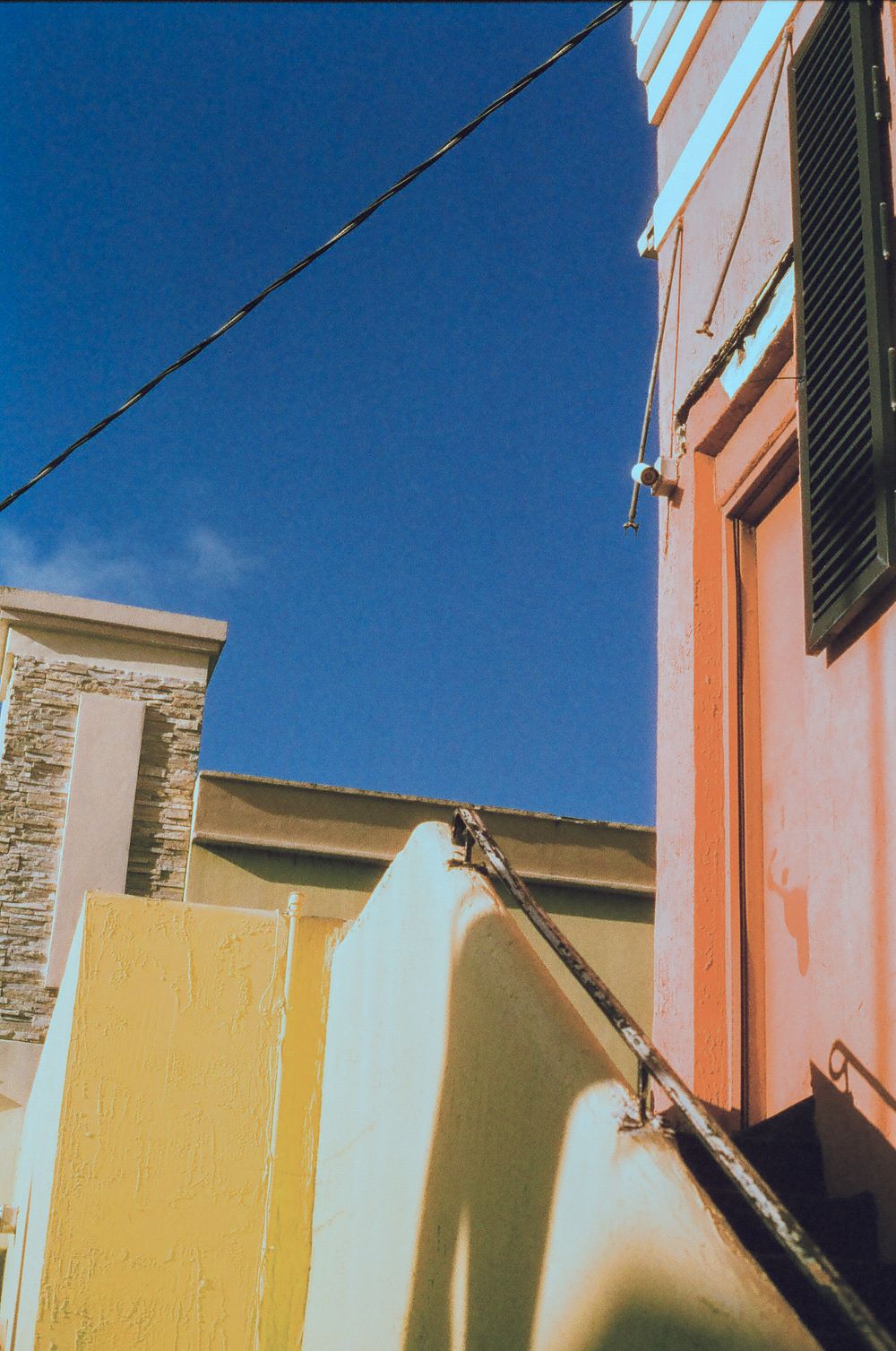Colorful Mediterranean buildings with yellow and pink walls under a clear blue sky, with power lines and shuttered windows.