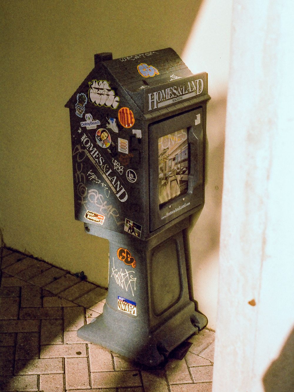 Vintage Homesland newspaper vending box covered in stickers and graffiti on a brick sidewalk.