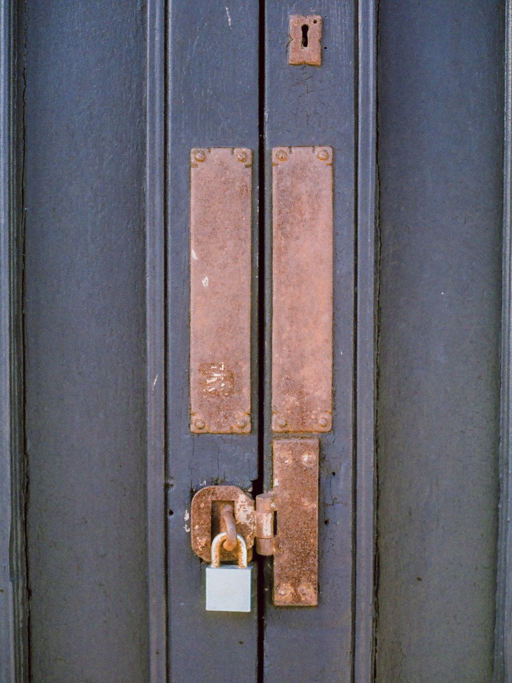A dark wooden door secured with a rusty padlock and aged copper push plates on a blue painted surface.