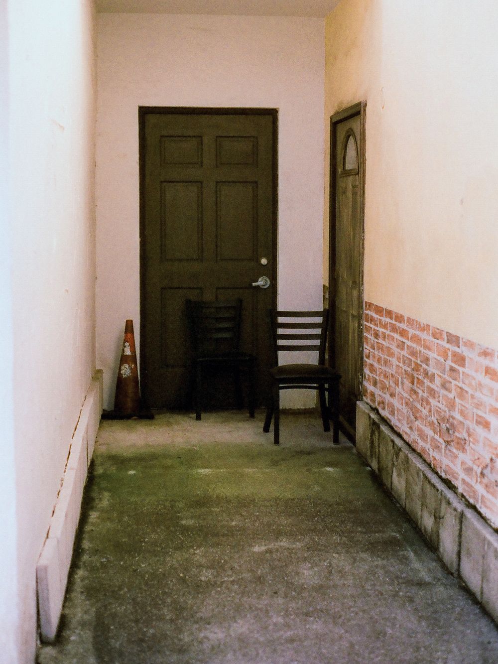 A narrow hallway with concrete floors, pink walls, brick accents, and two dark wooden chairs flanking a green door.