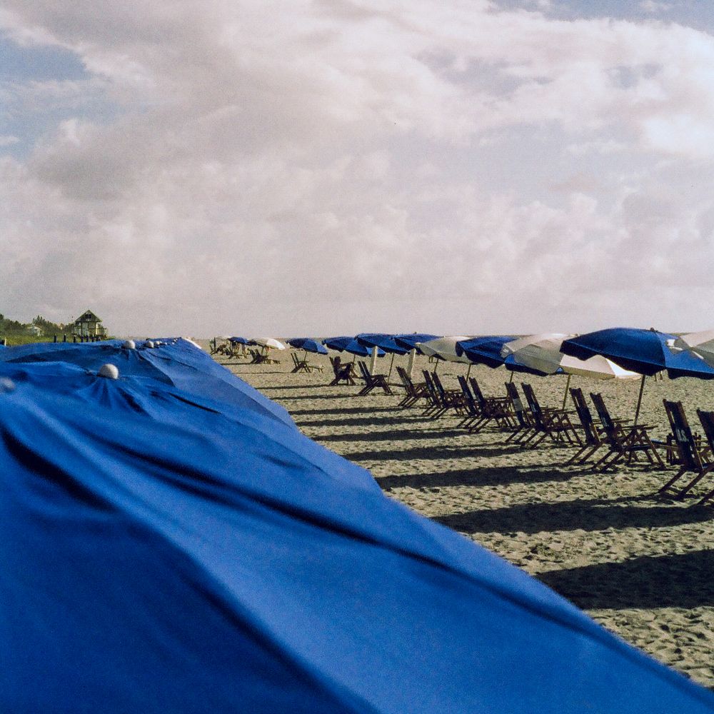 Rows of blue umbrellas and wooden lounge chairs line a sandy beach under a cloudy sky.