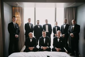 Groomsmen in black tuxedos stand together in a modern hotel room.
