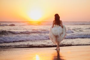 Person in white dress walking along beach shore during golden hour sunset.