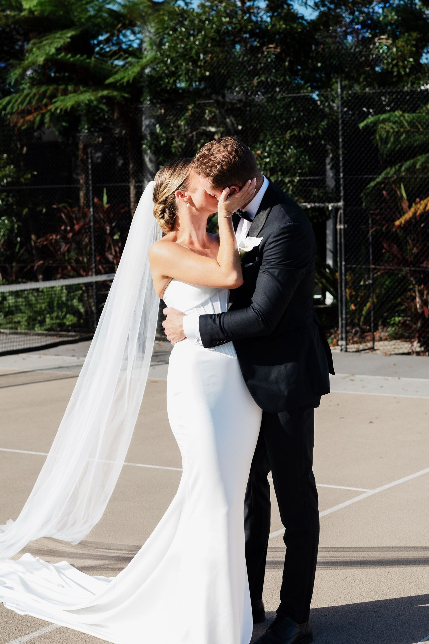 Bride and groom share a romantic moment at a tennis court net, bride wearing a flowing white gown and veil.