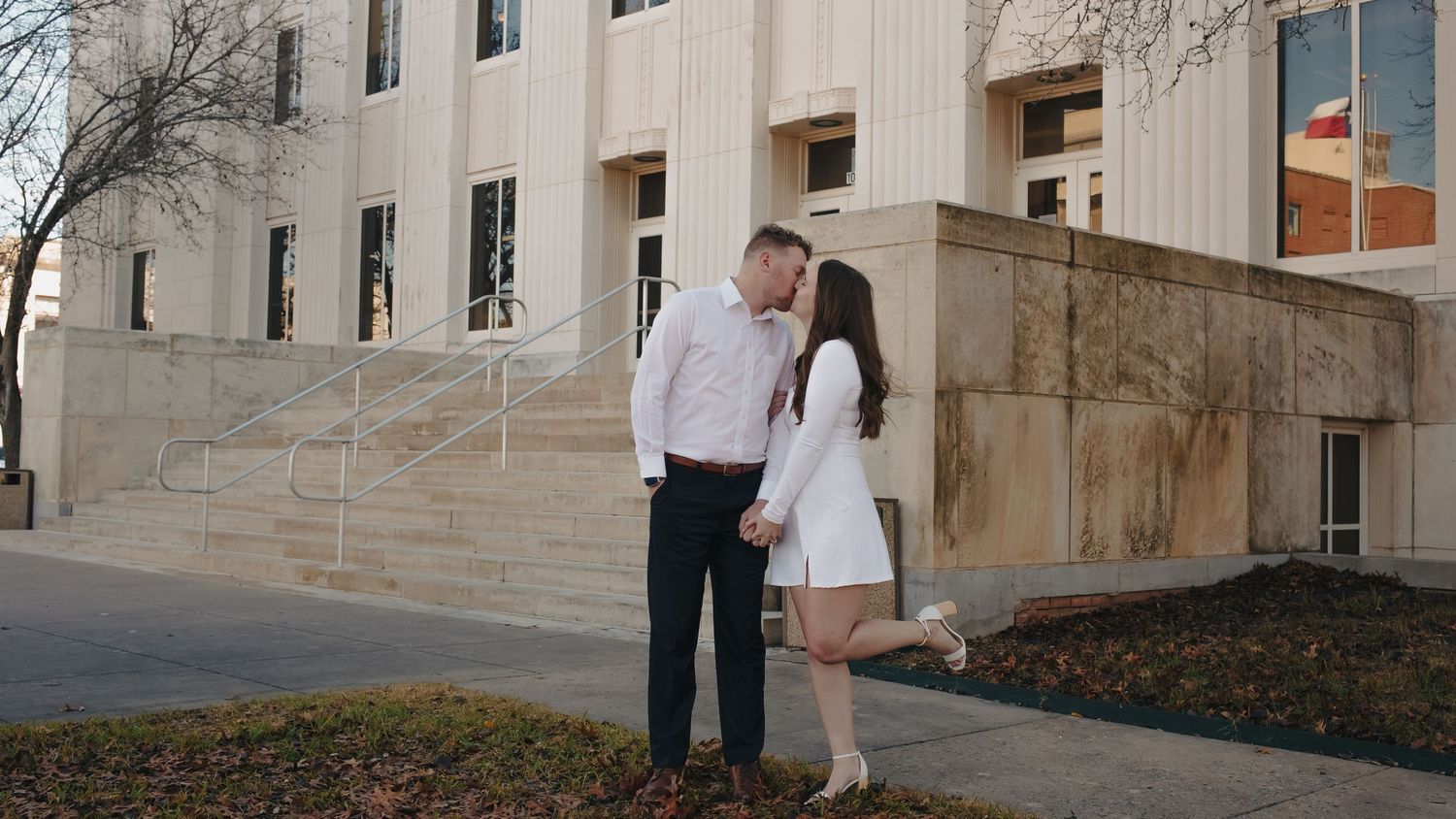 Couple sharing a romantic kiss outside a classic stone building, holding hands in a tender engagement photo.
