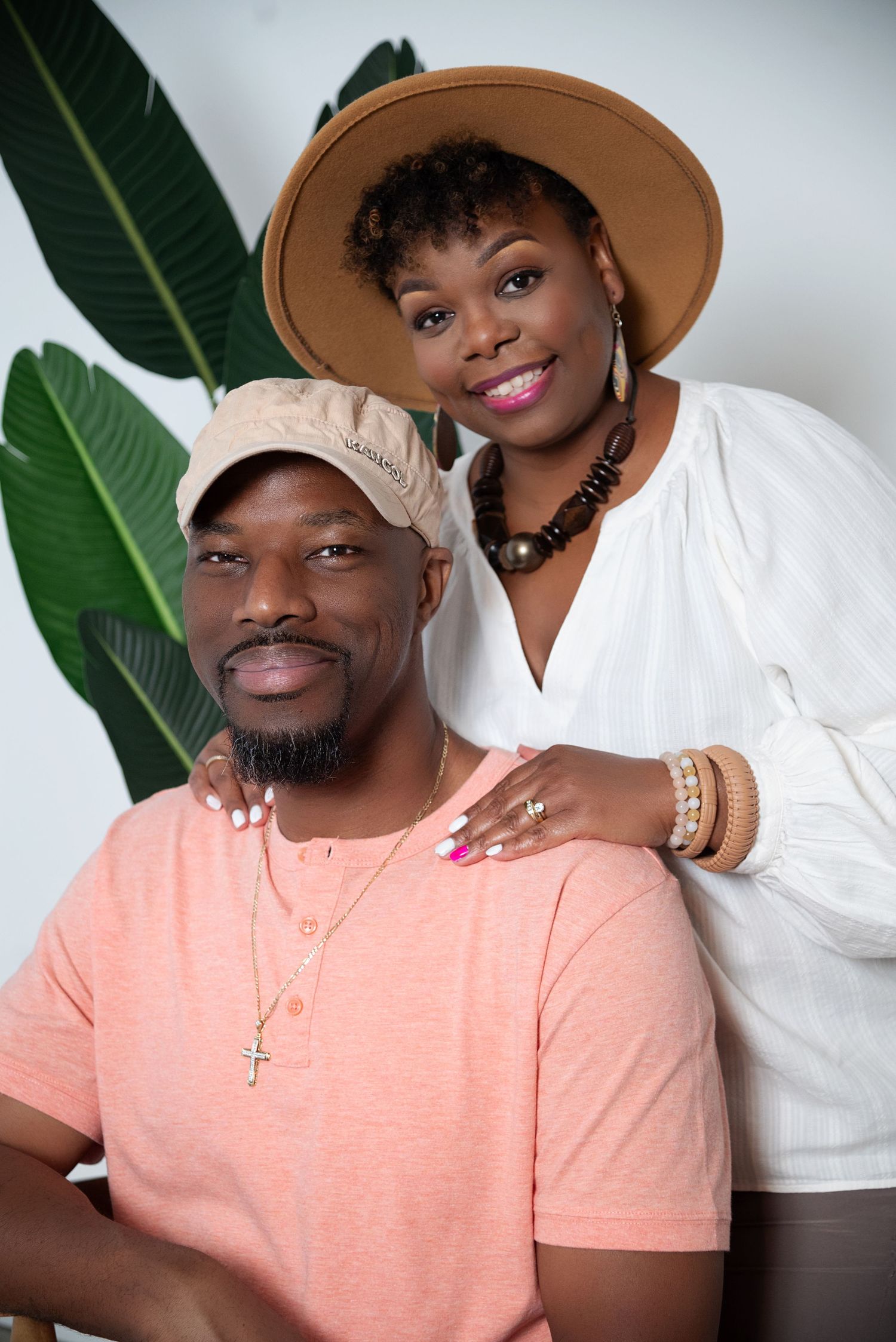 A couple in coordinating outfits of peach and white pose together against a white wall with tropical leaves.