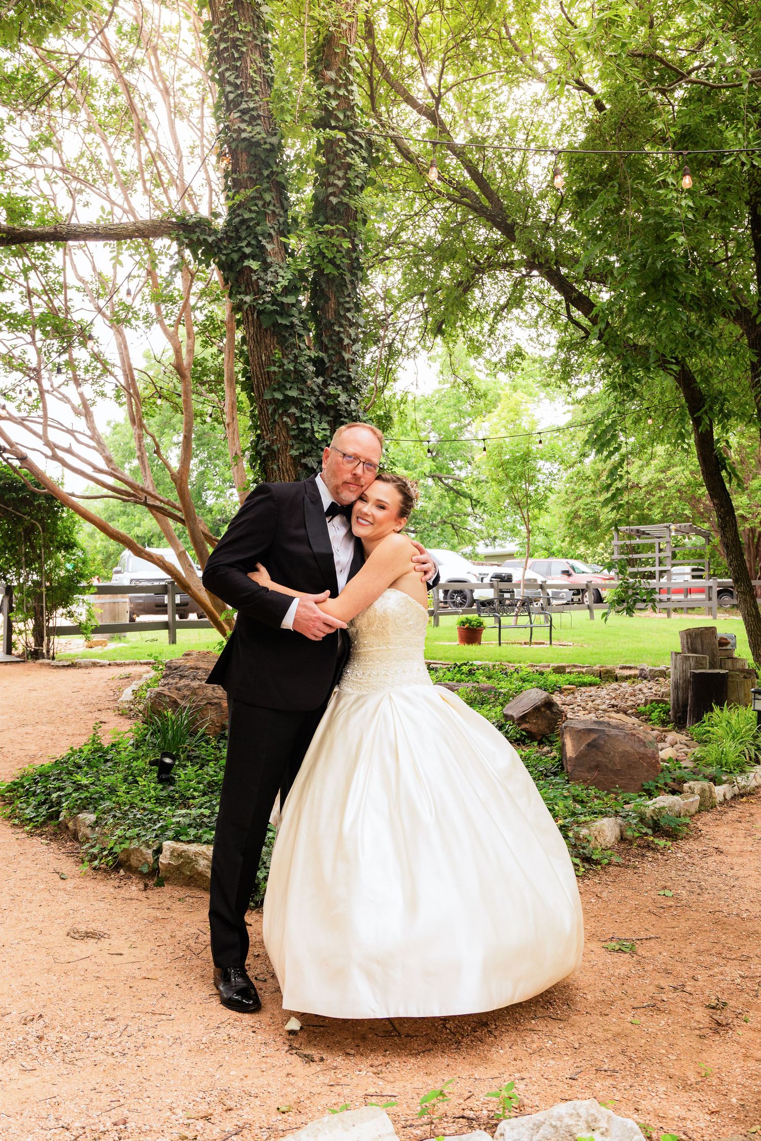 Bride in white ball gown embraces groom in black tuxedo outdoors among lush green trees on their wedding day.