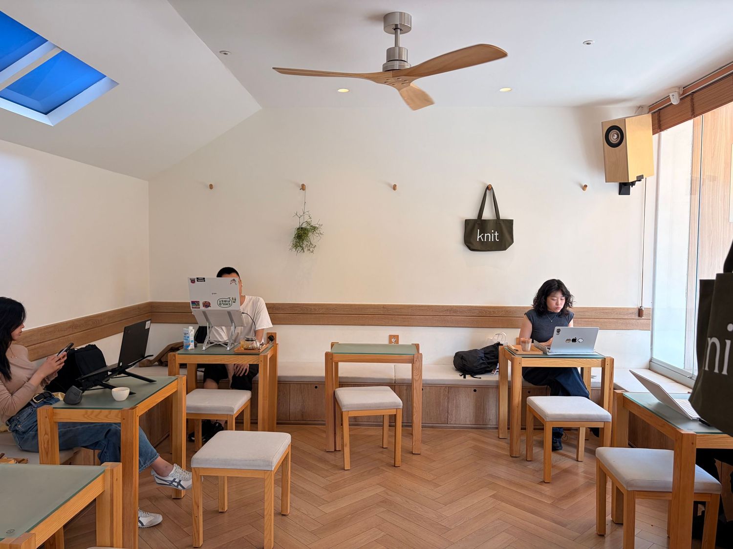 Cozy minimalist cafe interior with wooden furniture, ceiling fan, and patrons working on laptops in a bright, airy space.