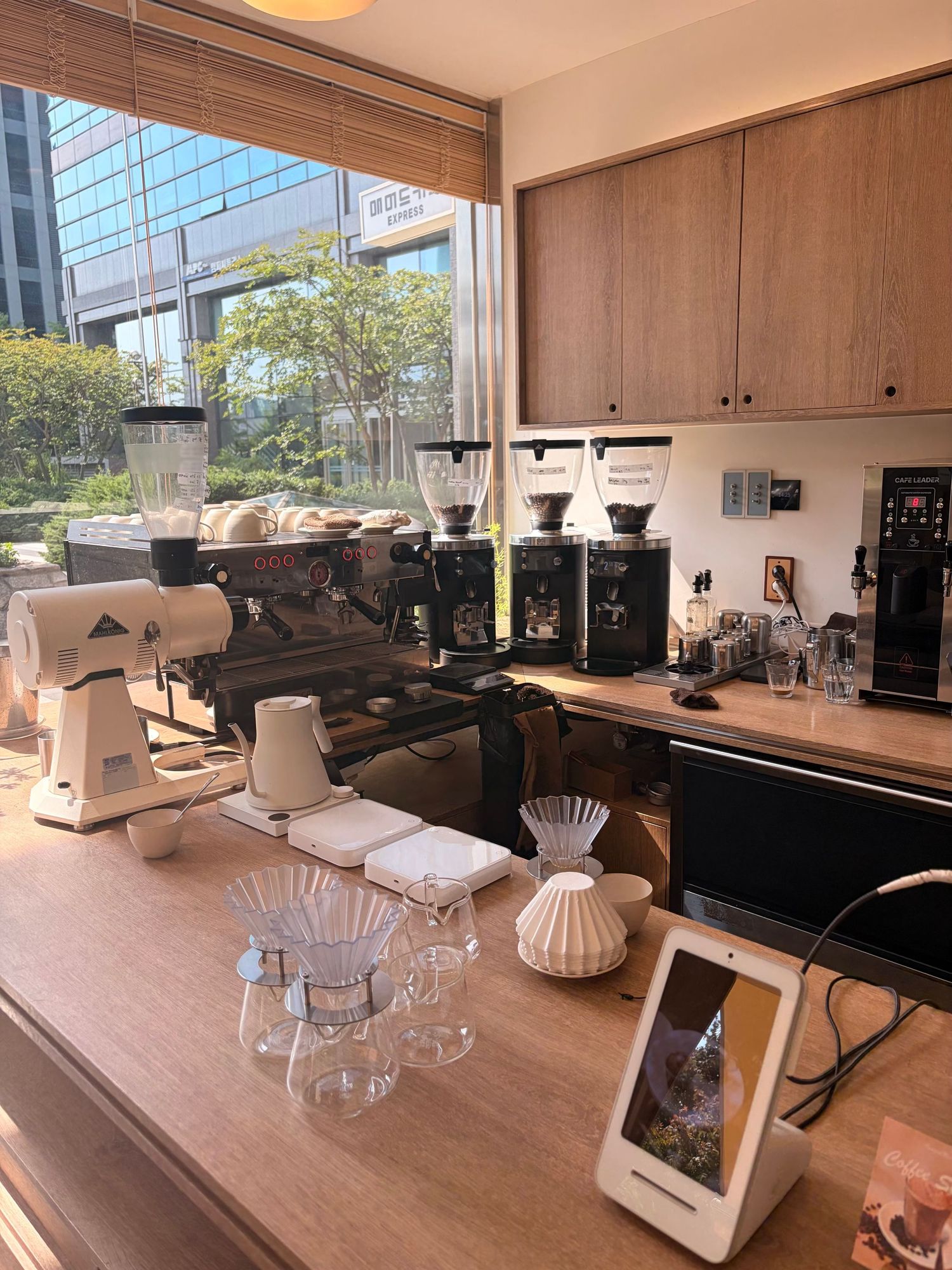 Modern coffee bar with espresso machines, grinders, Chemex brewers, and a tablet POS system on a wooden counter with city views.