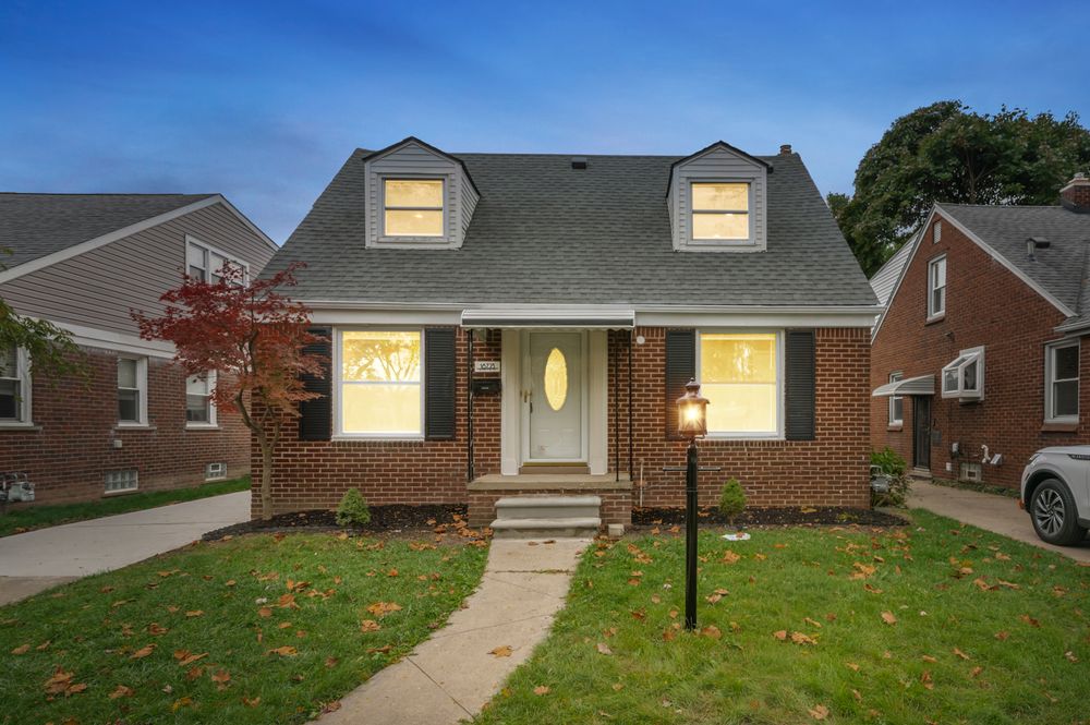 Charming brick Cape Cod home with dormer windows, manicured lawn, and warm interior lighting at dusk.