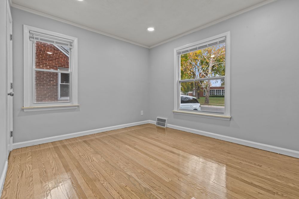 Bright empty room with hardwood floors, gray walls, crown molding, and two white-trimmed windows with natural light.